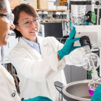 Two female scientists in whitecoats, gloves and goggles inspect a same from a machine.